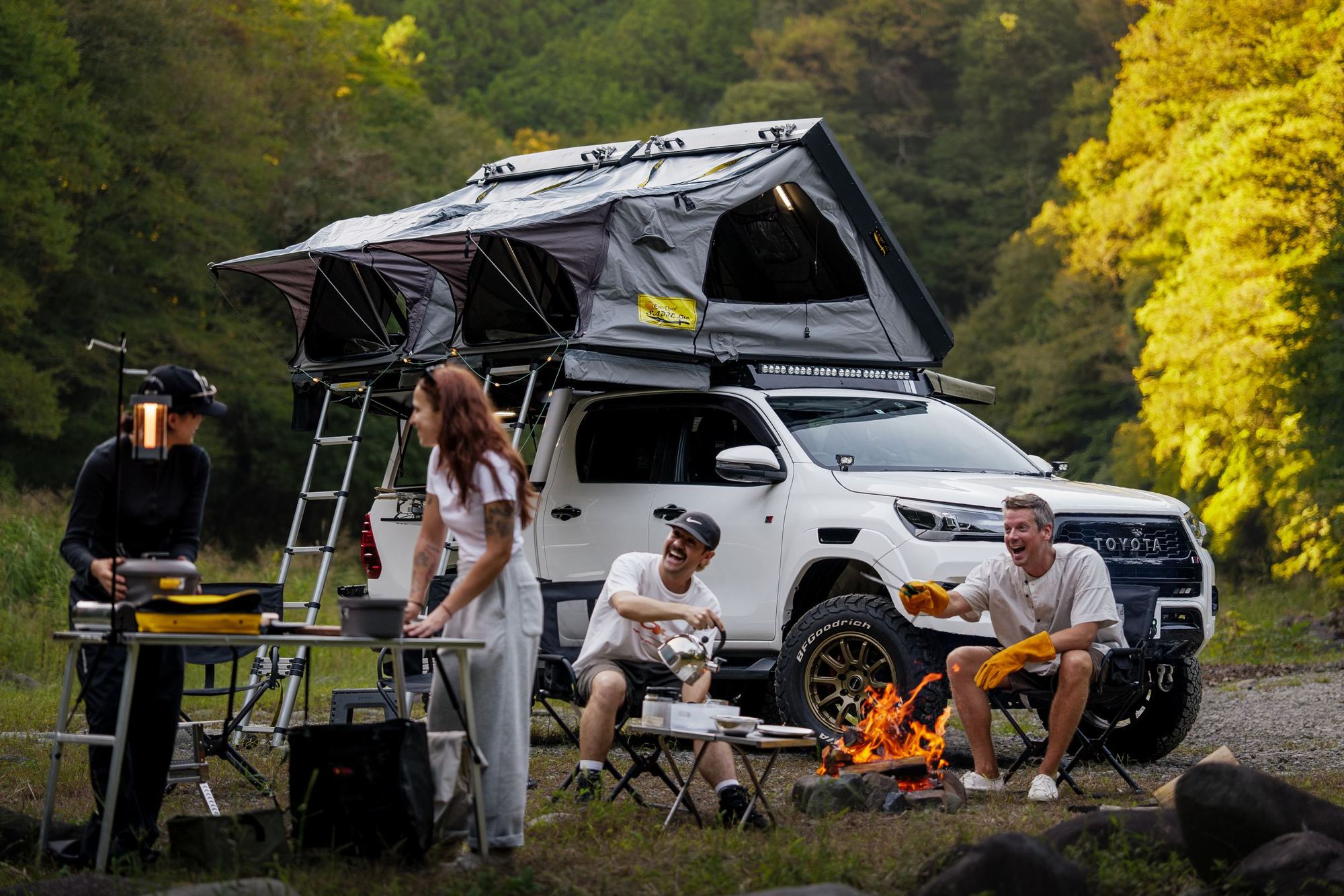 Friends laughing round the fire at camp, kettle on, with a rooftop tent open on a white Toyota Hilux.